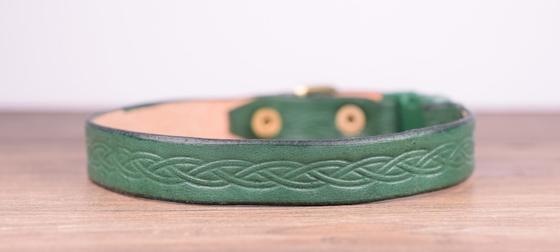 May include: A green leather bracelet featuring a Celtic knot pattern. The bracelet has a brass buckle and is approximately 2 cm wide. The inside of the bracelet is a natural tan colour.