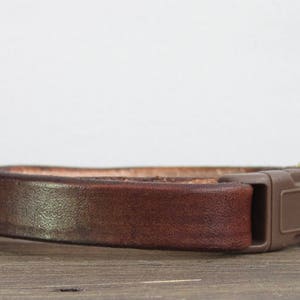 May include: A brown leather cat collar with a gold buckle and a brown plastic buckle. The collar has a small gold bell attached.