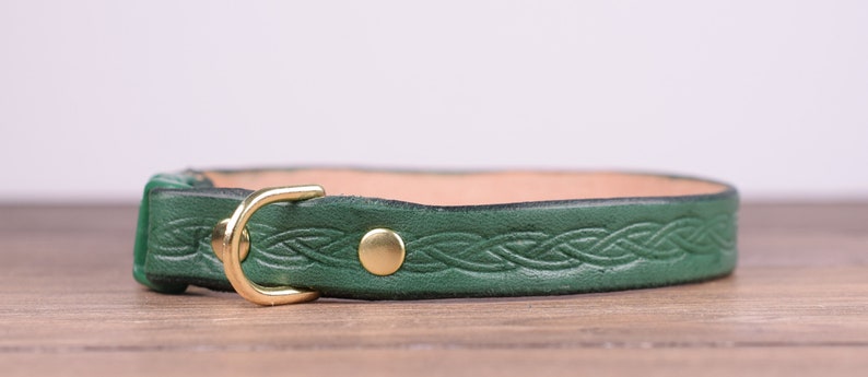 May include: A green leather collar with a brass buckle and a brass rivet. The collar features a Celtic knot design and is displayed on a wooden surface. The collar is approximately 2.5 cm wide.