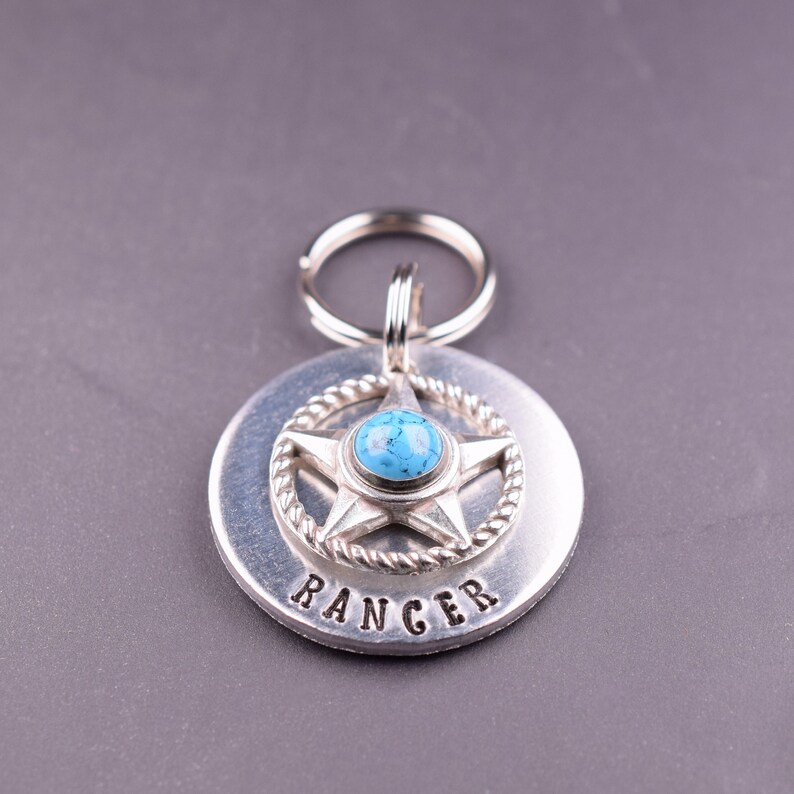 Puede incluir: Un llavero plateado con una etiqueta redonda y un colgante en forma de estrella. El colgante presenta una piedra turquesa en el centro de una estrella, rodeada por un dise&ntilde;o de cuerda. La palabra "RANGER" est&aacute; estampada en la etiqueta.