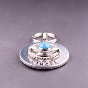 Puede incluir: Colgante plateado con una piedra turquesa engastada en un dise&ntilde;o de estrella, rodeada por un borde en forma de cuerda. La palabra "RANGER" est&aacute; estampada en la base circular. Un anillo plateado est&aacute; unido en la parte superior.