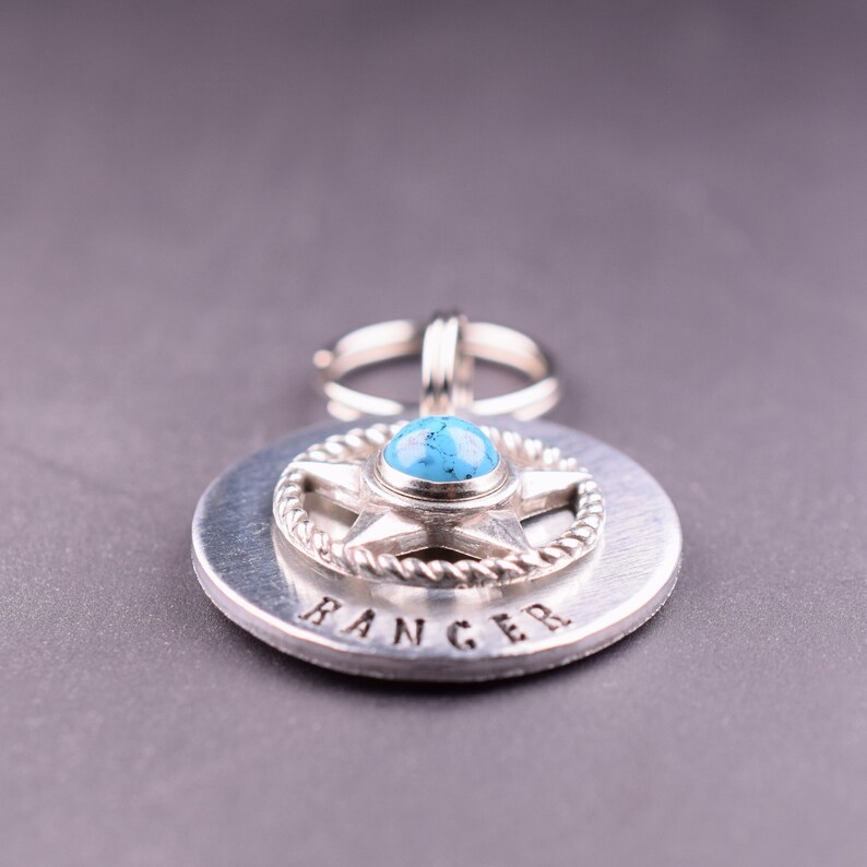 Puede incluir: Colgante plateado con una piedra turquesa engastada en un dise&ntilde;o de estrella, rodeada por un borde en forma de cuerda. La palabra "RANGER" est&aacute; estampada en la base circular. Un anillo plateado est&aacute; unido en la parte superior.