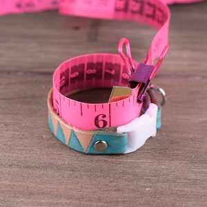 May include: A pink measuring tape is wrapped around a teal and tan leather bracelet with a white clasp. The tape is held in place with a purple binder clip. The bracelet has a geometric design.