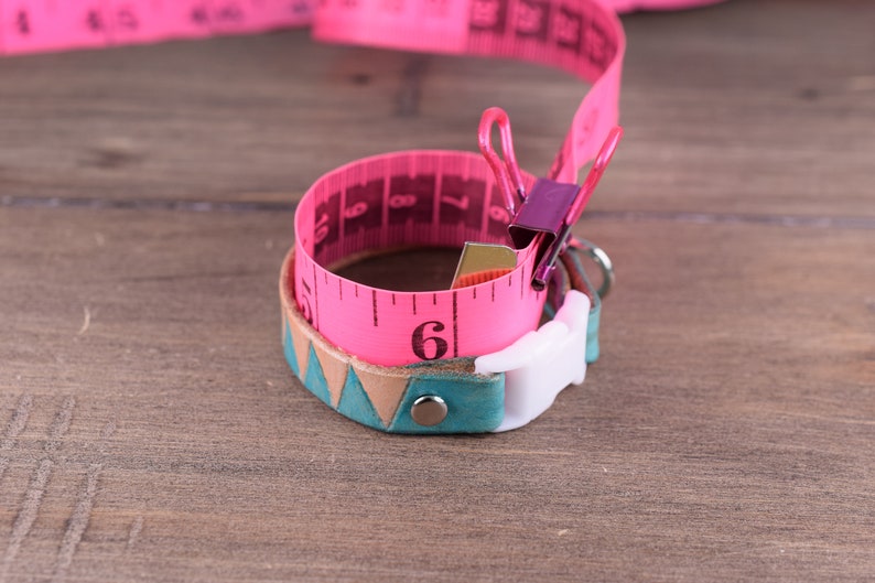 May include: A pink measuring tape is wrapped around a teal and tan leather bracelet with a white clasp. The tape is held in place with a purple binder clip. The bracelet has a geometric design.