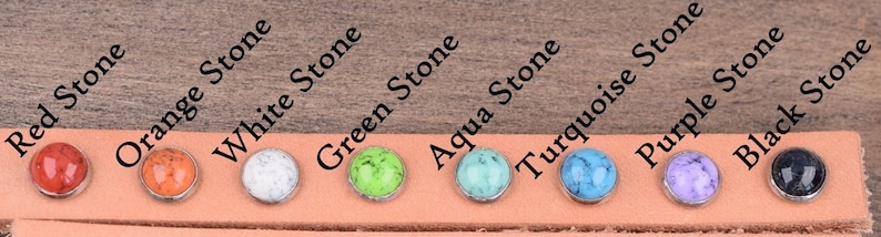 Puede incluir: Una selecci&oacute;n de cabujones de piedra de colores: rojo, naranja, blanco, verde, turquesa, morado y negro, cada uno engastado en un bisel plateado. Las piedras se muestran sobre una tira de cuero marr&oacute;n claro, con los nombres de las piedras impresos.