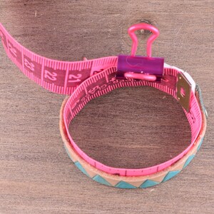 May include: Pink measuring tape wrapped around a brown leather bracelet with a turquoise and brown pattern. The tape is held together with a purple binder clip.