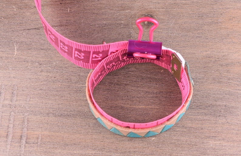 May include: Pink measuring tape wrapped around a brown leather bracelet with a turquoise and brown pattern. The tape is held together with a purple binder clip.