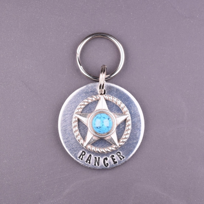 Puede incluir: Una placa para mascotas de color plateado con un dise&ntilde;o de estrella y la palabra "RANGER" grabada. La estrella tiene un centro turquesa y un borde en forma de cuerda. Un llavero plateado est&aacute; adjunto en la parte superior. Di&aacute;metro: aprox. 3,8 cm.