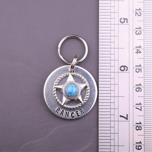 Puede incluir: Colgante de llavero plateado con una piedra turquesa engastada en un dise&ntilde;o de estrella, rodeada por un borde en forma de cuerda. La palabra "RANGER" est&aacute; estampada debajo de la estrella. El colgante est&aacute; unido a un llavero plateado. El colgante mide aproximadamente 5 cm de di&aacute;metro.