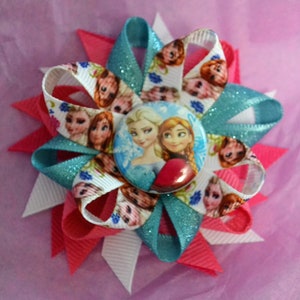 May include: A white, blue, and pink hair bow with a round button featuring the characters Elsa and Anna from the movie Frozen. The button has a white background with snowflakes and the characters are wearing their signature outfits.