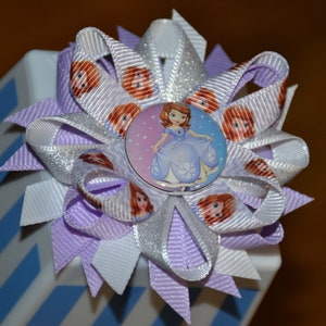 May include: A purple and white hair bow with a silver glitter ribbon and a center featuring a cartoon princess in a blue dress.