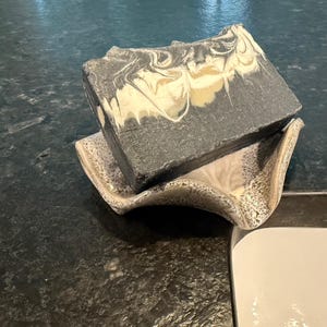 May include: A rectangular bar of handmade soap with a marbled design in shades of gray, white, and tan, resting on a decorative ceramic soap dish. The soap dish has a speckled pattern.
