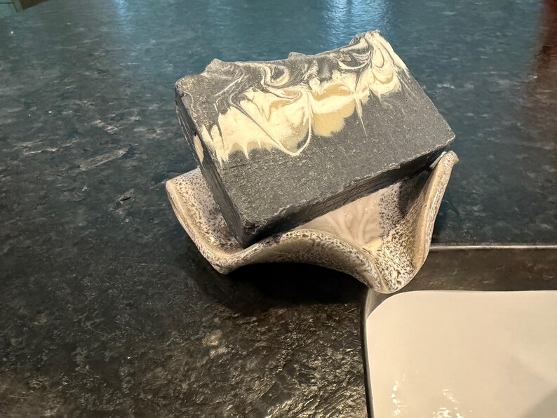 May include: A rectangular bar of handmade soap with a marbled design in shades of gray, white, and tan, resting on a decorative ceramic soap dish. The soap dish has a speckled pattern.
