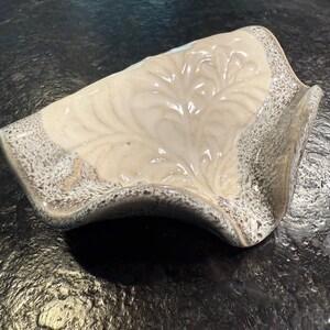 May include: A ceramic soap dish with a cream-colored interior featuring a leaf-like design. The dish has a speckled gray border and a curved, organic shape. The dish is on a dark surface.