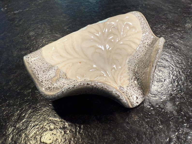 May include: A ceramic soap dish with a cream-colored interior featuring a leaf-like design. The dish has a speckled gray border and a curved, organic shape. The dish is on a dark surface.