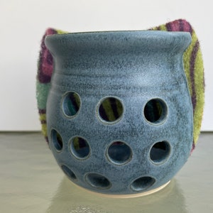 May include: A blue ceramic garlic keeper with multiple circular holes. A colorful, patterned cloth is partially visible behind the keeper. The keeper has a rounded shape and a wide opening at the top.