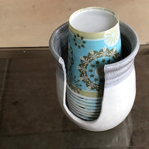 May include: A ceramic cup holder with a white and gray glaze, holding a stack of paper cups. The cups have a teal and brown floral pattern. The holder has a curved opening for easy access to the cups.