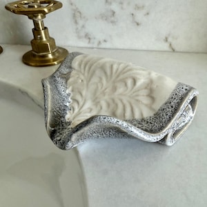 May include: A ceramic soap dish with a leaf design. The dish is off-white with a gray speckled border. The dish is near a brass faucet and sink.