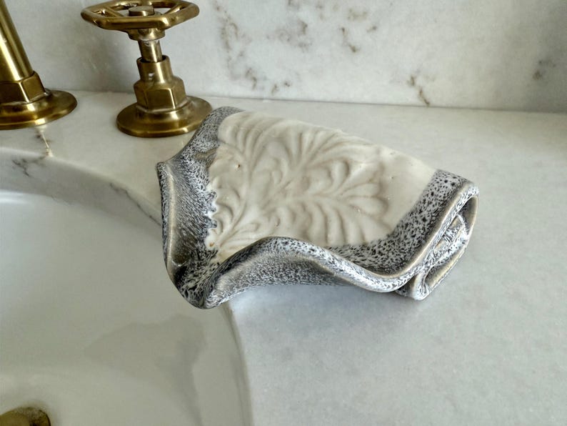 May include: A ceramic soap dish with a leaf design. The dish is off-white with a gray speckled border. The dish is near a brass faucet and sink.