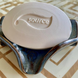 May include: A light beige bar of soap with the word "source" and a leaf design embossed on it, resting on a blue and brown ceramic soap dish. The dish has a unique, curved shape.