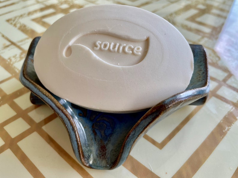 May include: A light beige bar of soap with the word "source" and a leaf design embossed on it, resting on a blue and brown ceramic soap dish. The dish has a unique, curved shape.