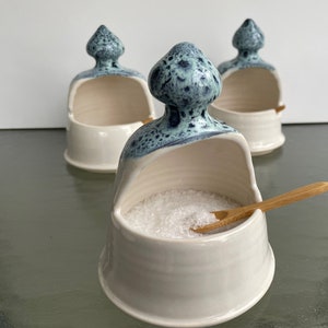 May include: Three white ceramic salt cellars with blue and white speckled glaze. The salt cellars have a unique design with a small opening at the top for pouring salt. One of the salt cellars is filled with salt and has a small wooden spoon.
