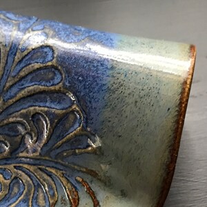 May include: Close-up of a ceramic piece with a blue and tan floral design. The piece has a glossy finish and a brown rim. The blue glaze fades into a lighter, almost white color. The floral pattern is raised and textured.