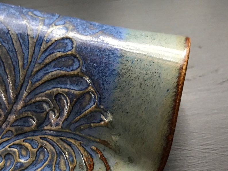 May include: Close-up of a ceramic piece with a blue and tan floral design. The piece has a glossy finish and a brown rim. The blue glaze fades into a lighter, almost white color. The floral pattern is raised and textured.