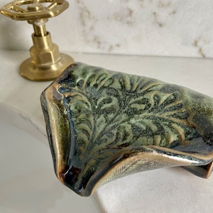 May include: A ceramic soap dish with a leaf pattern in shades of green and brown. The dish has a curved shape and a glossy finish. The edges are trimmed with a gold color. Bathroom decor.