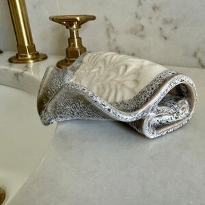 May include: A rolled, decorative ceramic towel in shades of cream, gray, and black. The towel has a speckled border and a textured, floral-like design on the top surface. The towel is resting on a white countertop near a sink with gold fixtures.