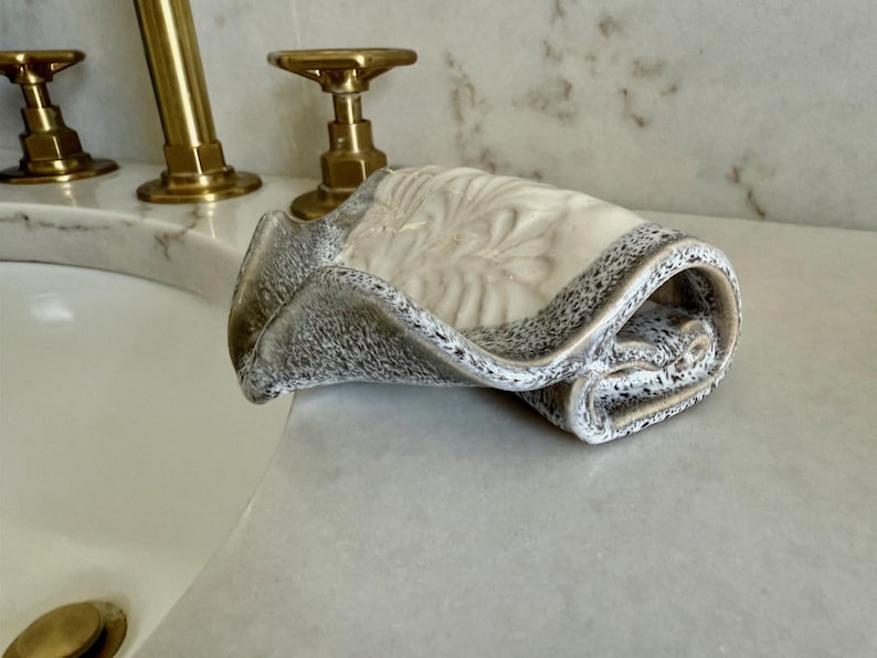 May include: A rolled, decorative ceramic towel in shades of cream, gray, and black. The towel has a speckled border and a textured, floral-like design on the top surface. The towel is resting on a white countertop near a sink with gold fixtures.