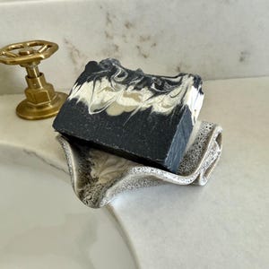 May include: A bar of soap with a black and white marbled design rests on a speckled ceramic soap dish. The soap dish is a light beige color and has a wavy design. The background includes a white countertop and brass fixtures.