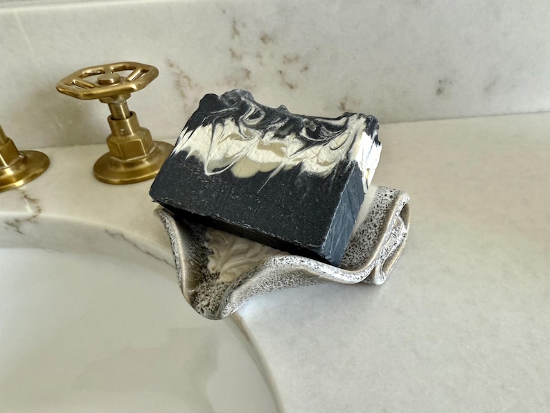 May include: A bar of soap with a black and white marbled design rests on a speckled ceramic soap dish. The soap dish is a light beige color and has a wavy design. The background includes a white countertop and brass fixtures.
