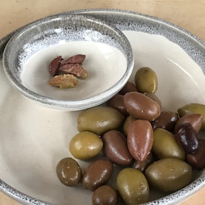 May include: A white ceramic serving dish with a smaller, attached bowl, both with a gray rim. The dish contains a variety of olives, including green and brown varieties. A small portion of dried olives is in the smaller bowl.