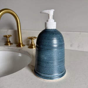 May include: A blue ceramic soap dispenser with a white pump top. The dispenser has a rounded shape and sits on a white countertop next to a gold faucet and sink.