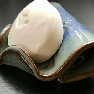 May include: A handmade ceramic soap dish with a wavy design, glazed in shades of blue and green with brown edging. A white bar of soap rests in the center. The dish is approximately 6 inches long.
