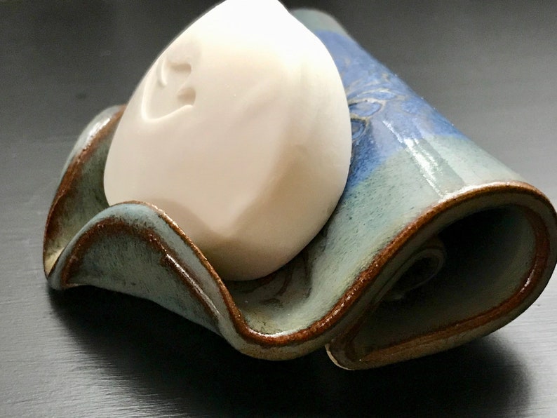 May include: A handmade ceramic soap dish with a wavy design, glazed in shades of blue and green with brown edging. A white bar of soap rests in the center. The dish is approximately 6 inches long.