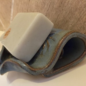 May include: A light-colored bar of soap rests on a blue ceramic soap dish. The dish has a wavy design with brown trim. The soap has a small orange stain. The soap dish is on a white surface.