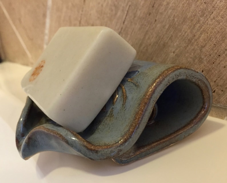 May include: A light-colored bar of soap rests on a blue ceramic soap dish. The dish has a wavy design with brown trim. The soap has a small orange stain. The soap dish is on a white surface.
