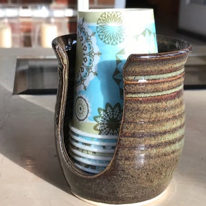 May include: A brown ceramic cup holder with a stack of paper cups inside. The cups have a blue and green floral pattern.