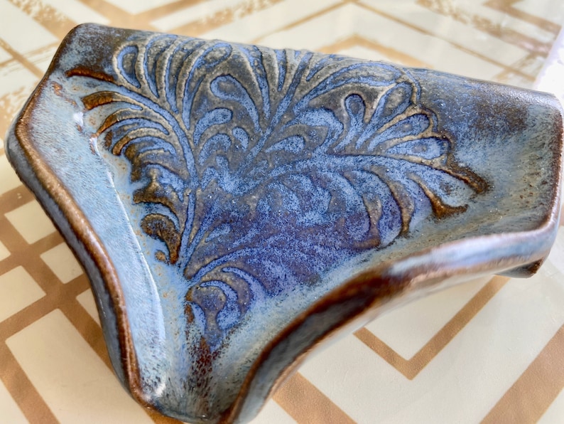 May include: A handcrafted ceramic soap dish with a blue and brown glaze. The dish features an embossed floral design in the center. The edges are curved and the dish has a glossy finish.