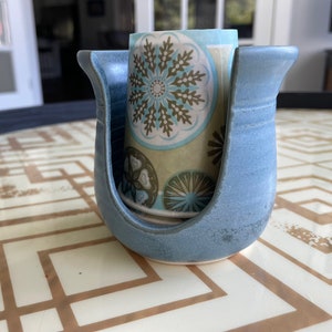 May include: A light blue ceramic napkin holder with a U-shaped design. The holder contains a stack of patterned napkins with a teal, brown, and white snowflake design. The holder sits on a table with a geometric pattern.