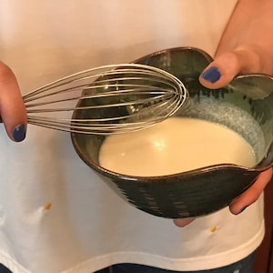 May include: A ceramic bowl with a dark green glaze holds a creamy white liquid. A stainless steel whisk is held above the bowl, ready to mix. The bowl has a unique, organic shape.