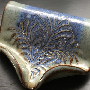 May include: Handmade ceramic soap dish with a blue and gray floral design. The dish has a curved shape with a raised edge and measures approximately 6 inches long and 4 inches wide. The dish is glazed with a glossy finish.