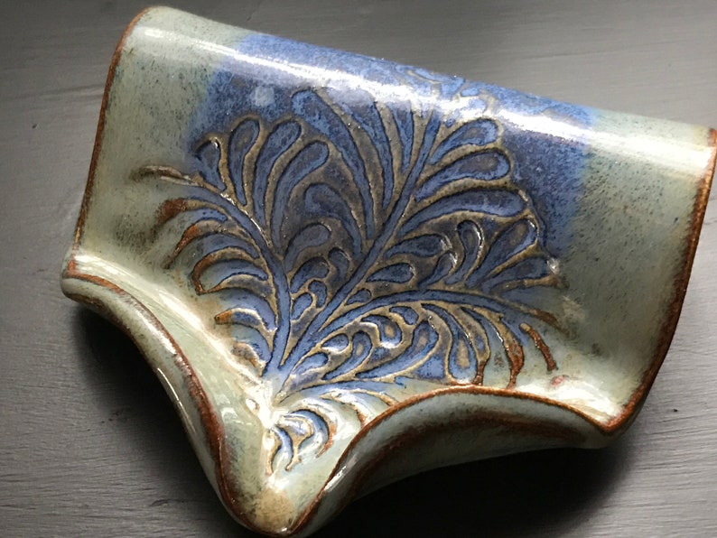 May include: Handmade ceramic soap dish with a blue and gray floral design. The dish has a curved shape with a raised edge and measures approximately 6 inches long and 4 inches wide. The dish is glazed with a glossy finish.
