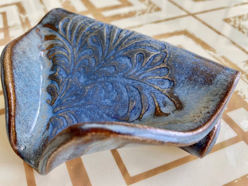 May include: A handmade ceramic soap dish with a blue glaze and a brown rim. The dish features an embossed floral design in the center. The edges are slightly curved, and the overall shape is rectangular.