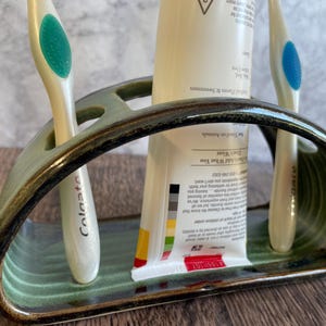 May include: A green ceramic toothbrush holder with two toothbrushes and a tube of toothpaste. The holder has a curved shape and a textured surface. The toothbrushes are white with blue bristles. The toothpaste tube is white with black and red text.