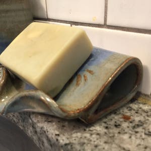 May include: A handmade ceramic soap dish with a bar of soap. The dish is a light blue with brown accents and a curved design. The soap is a light beige color. The dish is on a granite countertop.