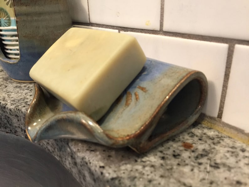 May include: A handmade ceramic soap dish with a bar of soap. The dish is a light blue with brown accents and a curved design. The soap is a light beige color. The dish is on a granite countertop.