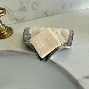 May include: A square bar of light-colored soap rests in a decorative soap dish. The dish has a gray and white floral design and a textured rim. A brass faucet is visible in the background, suggesting a bathroom setting.